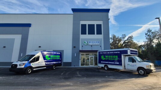 Two Mission Storage branded moving trucks parked strategically in front of the entrance to the Mission Storage Facility entrance